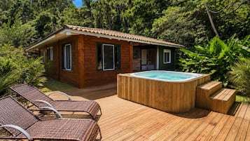 A wooden cabin with a hot tub and lounge chairs on a deck surrounded by lush greenery.