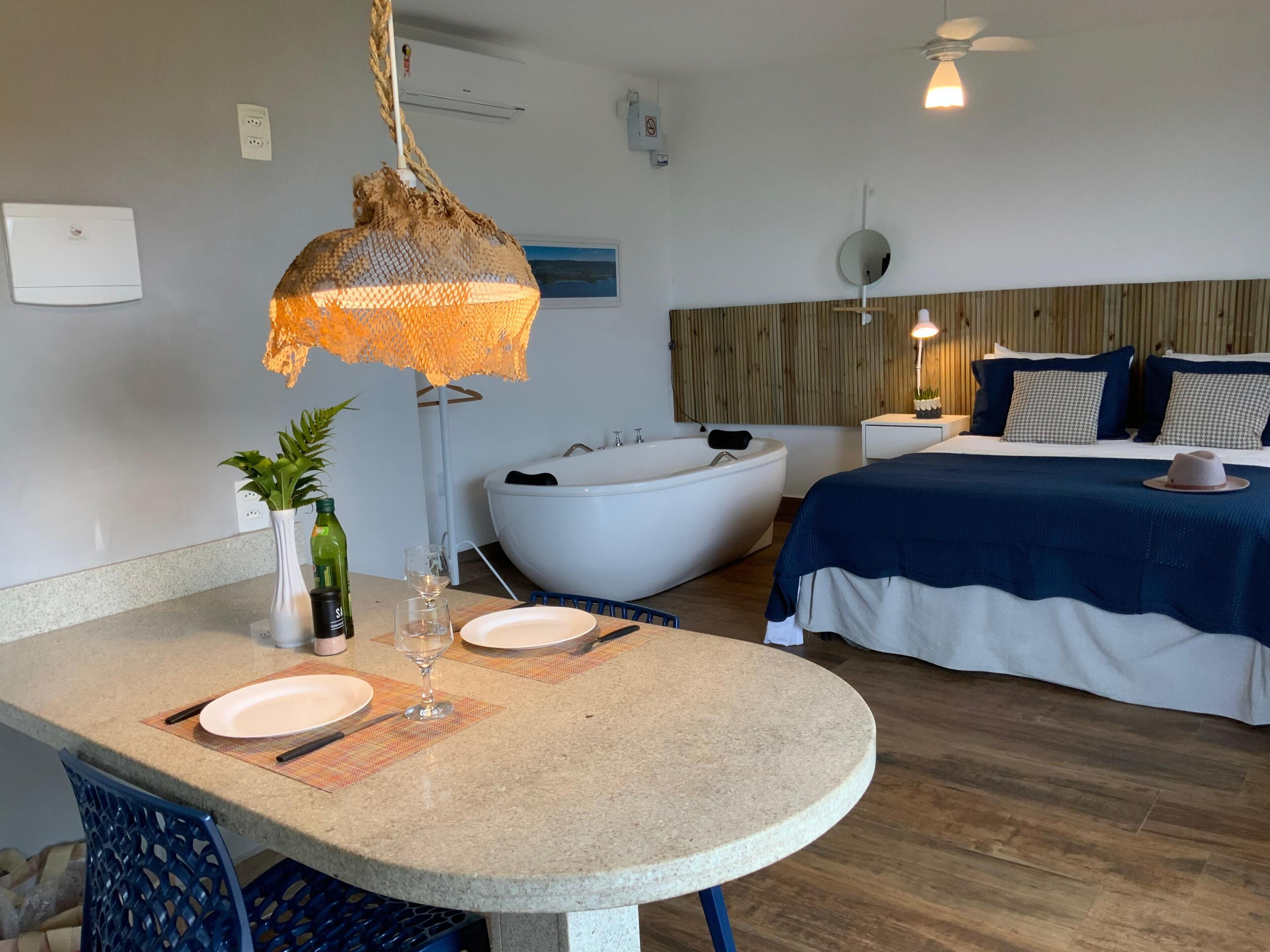 A modern hotel room with a round dining table, a large bathtub, a bed with a blue and white bedspread, and a hanging light fixture.