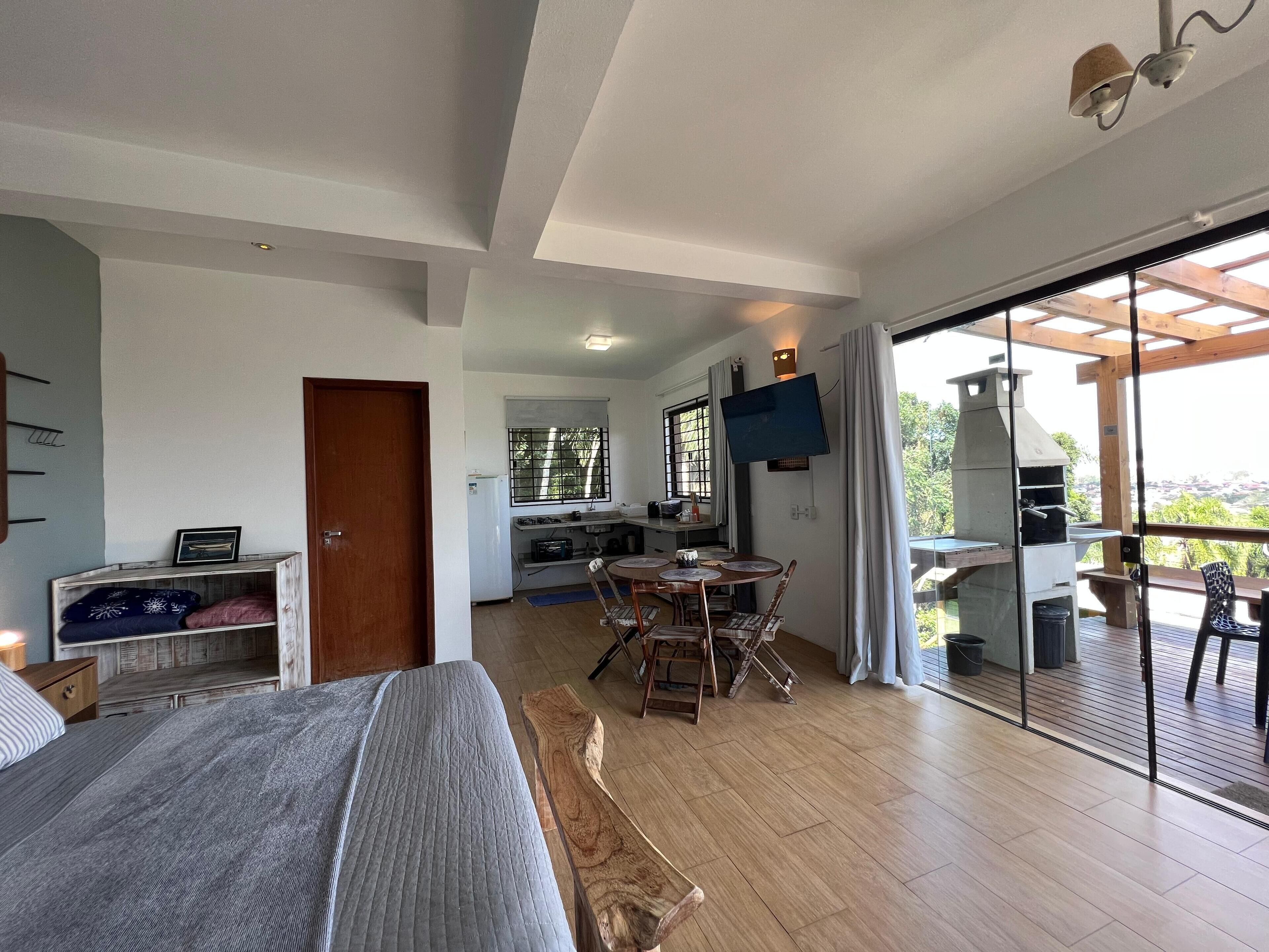 A modern bedroom with a large bed, a wooden nightstand, and a bedside table. There is a kitchen area with a dining table and chairs, and a balcony with a grill.