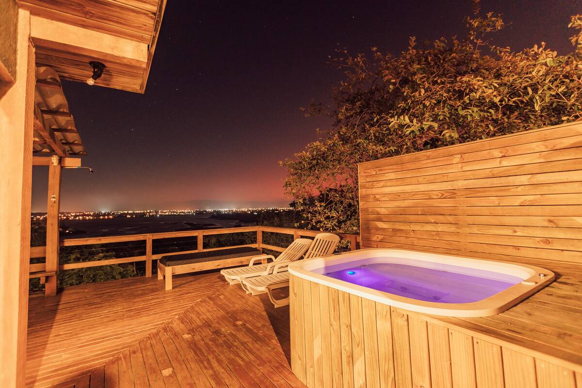 A wooden deck with a hot tub and lounge chairs, overlooking a cityscape at night.