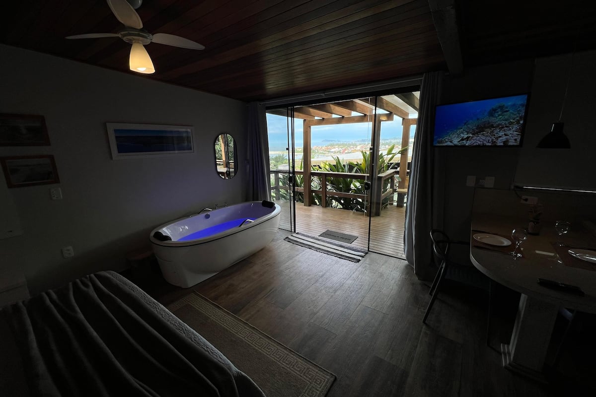 A modern bathroom with a freestanding bathtub, a ceiling fan, and a large window offering a view of the outdoors.