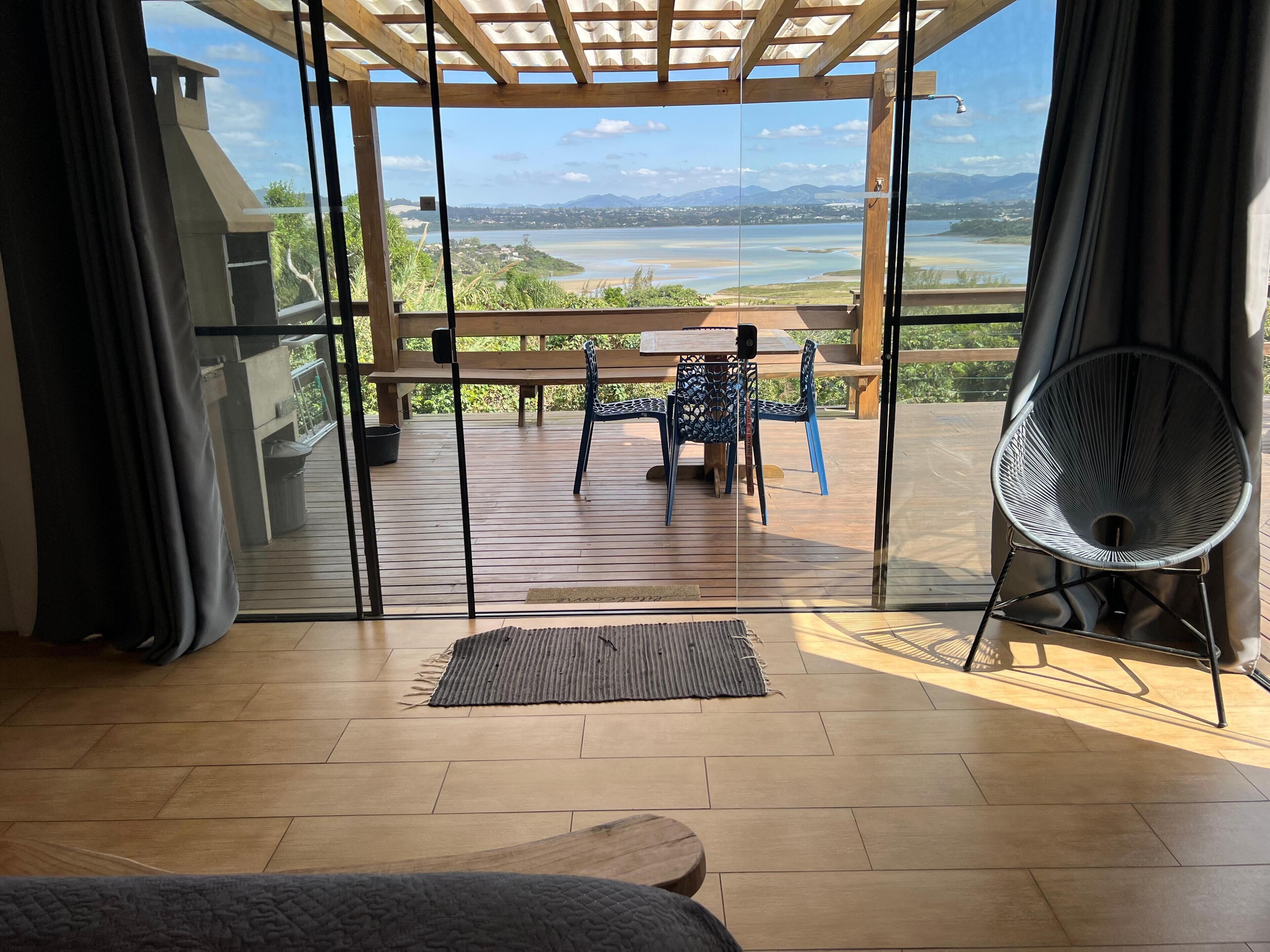 Una habitación con piso de madera, una cama, vista a una terraza de madera con mesa y sillas, y un ventilador.