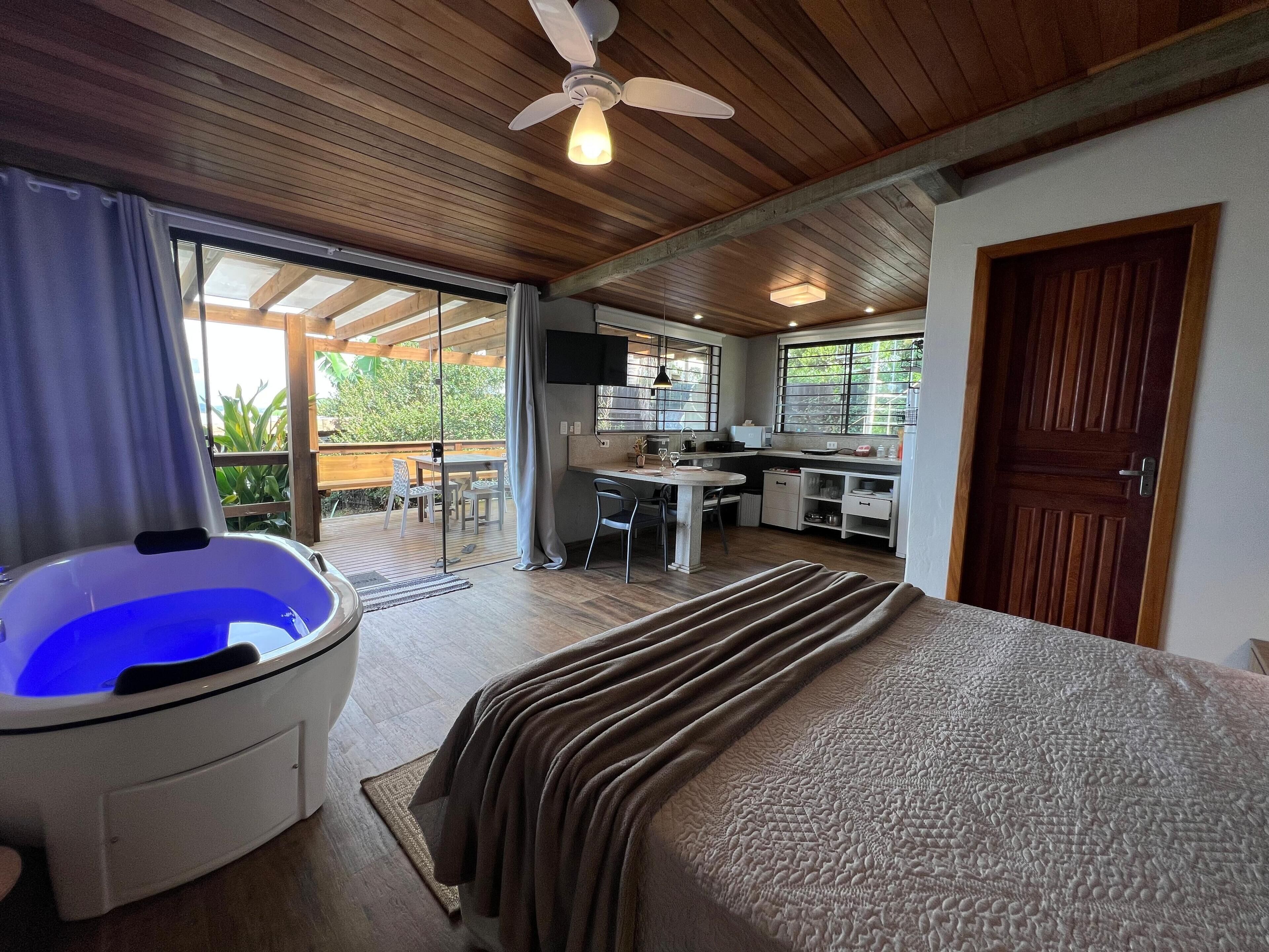 Un dormitorio con una cama grande, una bañera de hidromasaje, una kitchenette y un ventanal corredera a un patio.