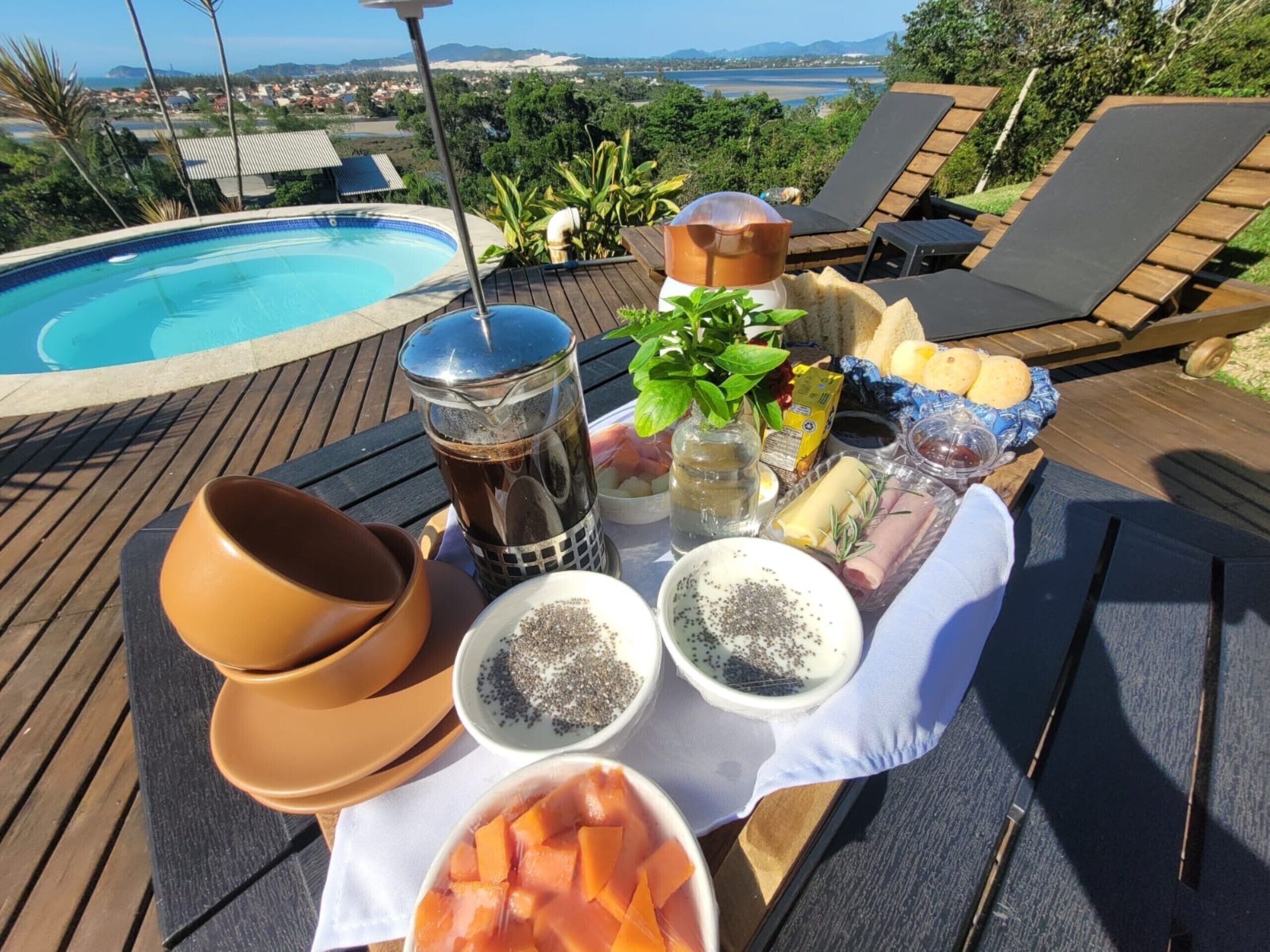 Mesa junto a la piscina con diversos alimentos, como queso, pan y una bebida, frente a una piscina y sillones de descanso.