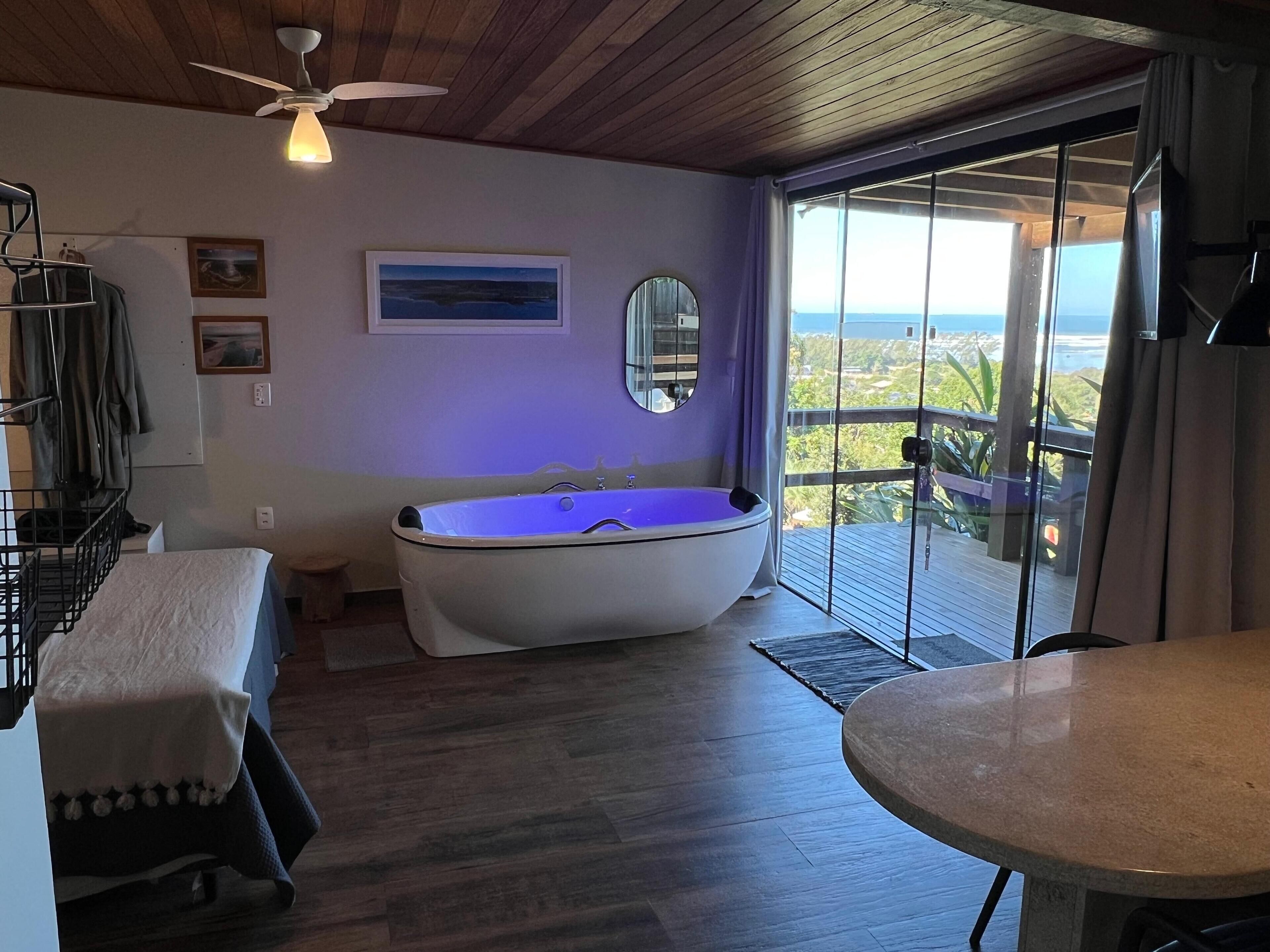 A room with a wooden ceiling, a ceiling fan, a bathtub, a table, and a balcony with a view.