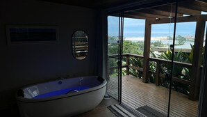 Une salle de bain moderne avec une baignoire sur pieds, un miroir et une vue sur un balcon doté d’une terrasse en bois et de verdure.