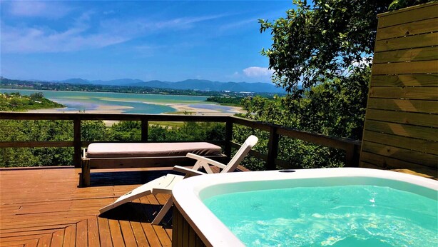 Une terrasse en bois avec un jacuzzi et des chaises longues, offrant une vue sur une étendue d’eau et des montagnes lointaines.