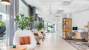 Un área de espera moderna con una fila de sofás y sillones blancos, un escritorio de madera con una computadora, y ventanas amplias que ofrecen vistas al exterior.