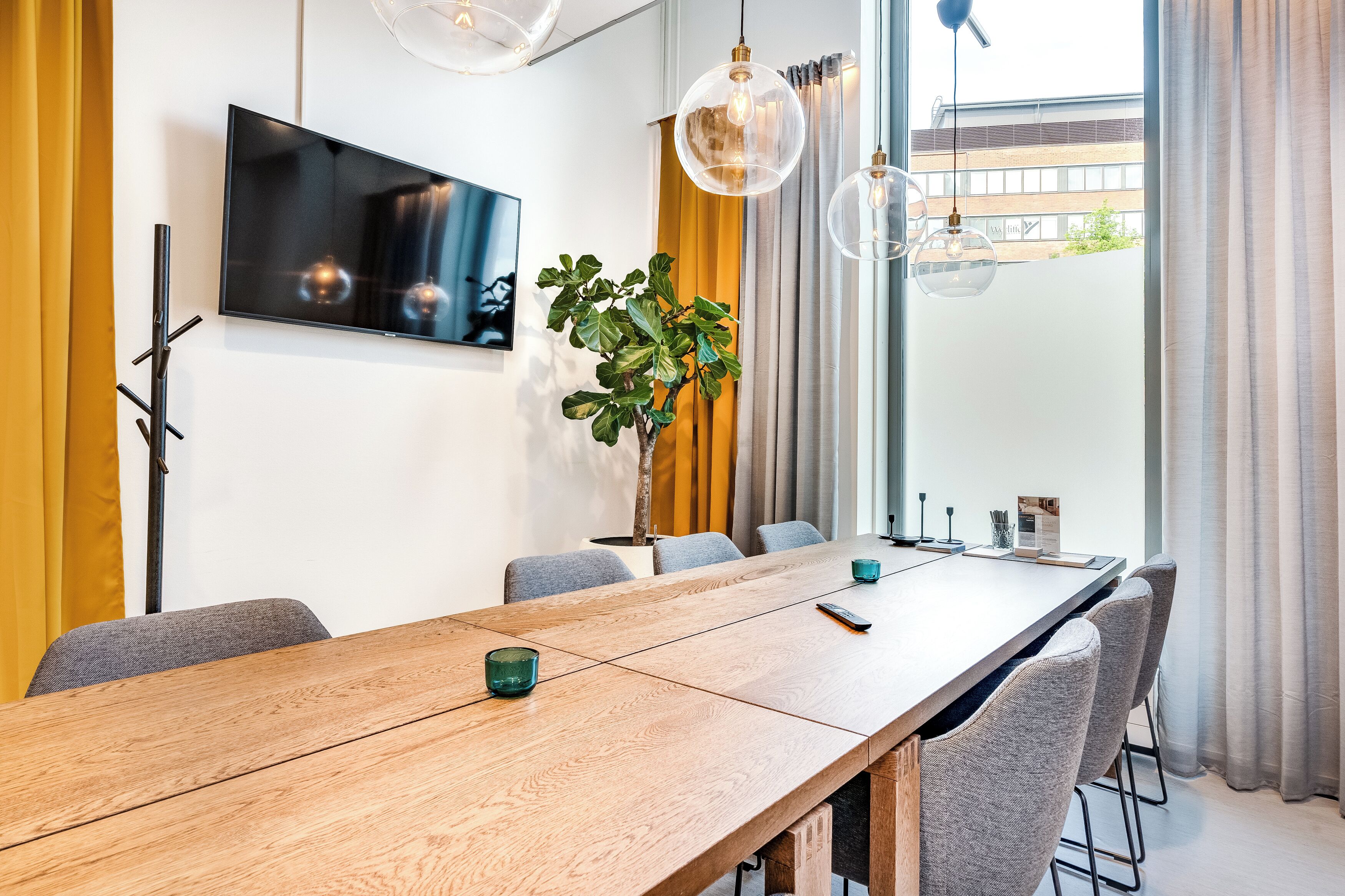 A modern dining area with a long wooden table, grey upholstered chairs, a potted plant, and a mounted flat-screen TV.