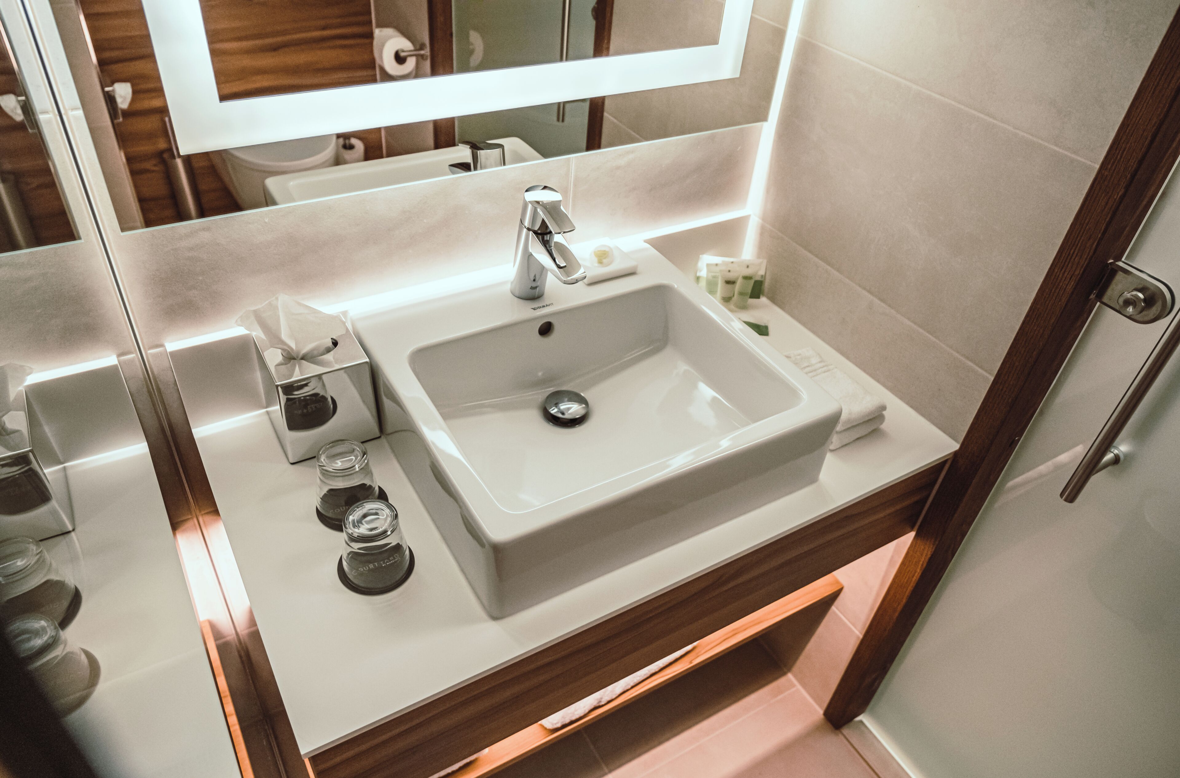 Un baño moderno con lavamanos blanco, espejo y tocador de madera.