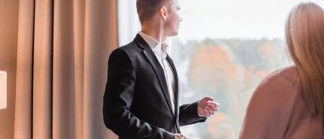 A man in a black suit standing by a window, conversing with a woman.
