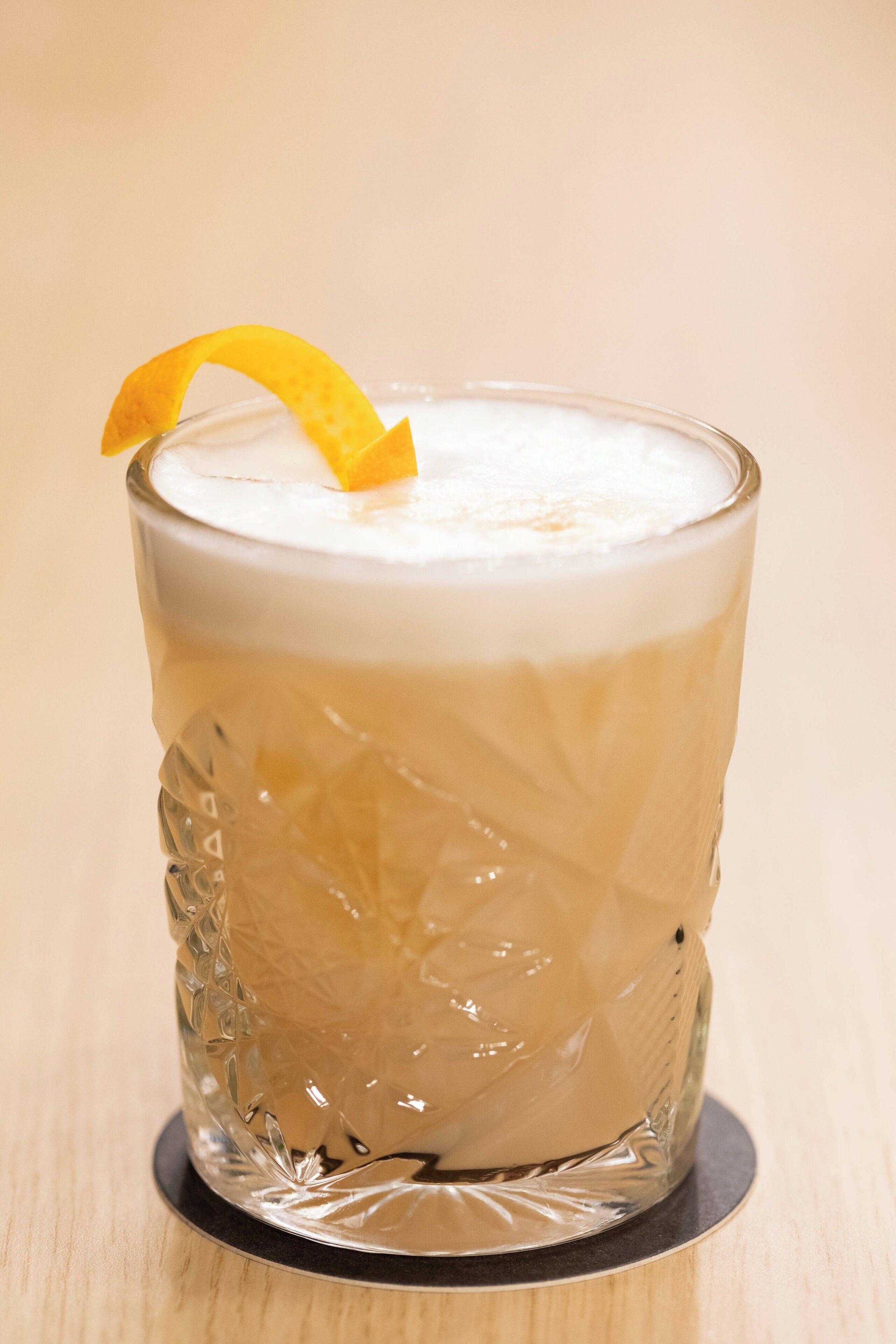 A clear glass with a textured pattern, a white foam layer, and a lemon peel garnish on a coaster.
