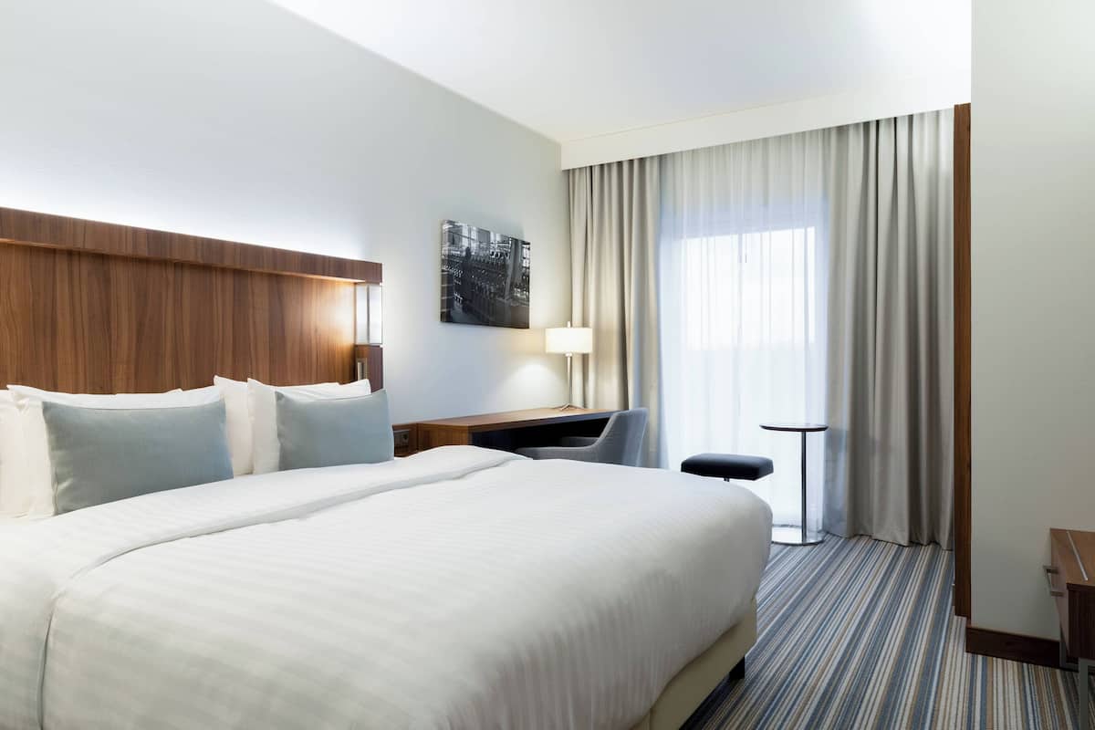 Hotellihuone, jossa on suuri sänky, työpöytä, tuoli, lamppu ja ikkunasta avautuva näkymä kaupunkiin.