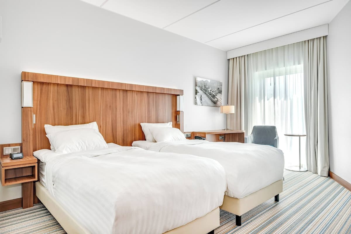 Hotellihuone, jossa on kaksi sänkyä, puinen pääty, yöpöytä lampulla, tuoli ja ikkuna, jossa on verhot.