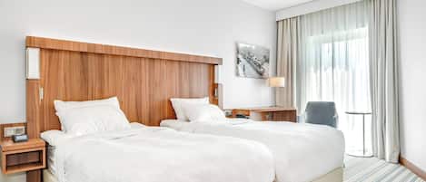A hotel room with two beds, a wooden headboard, a bedside table with a lamp, a chair, and a window with curtains.