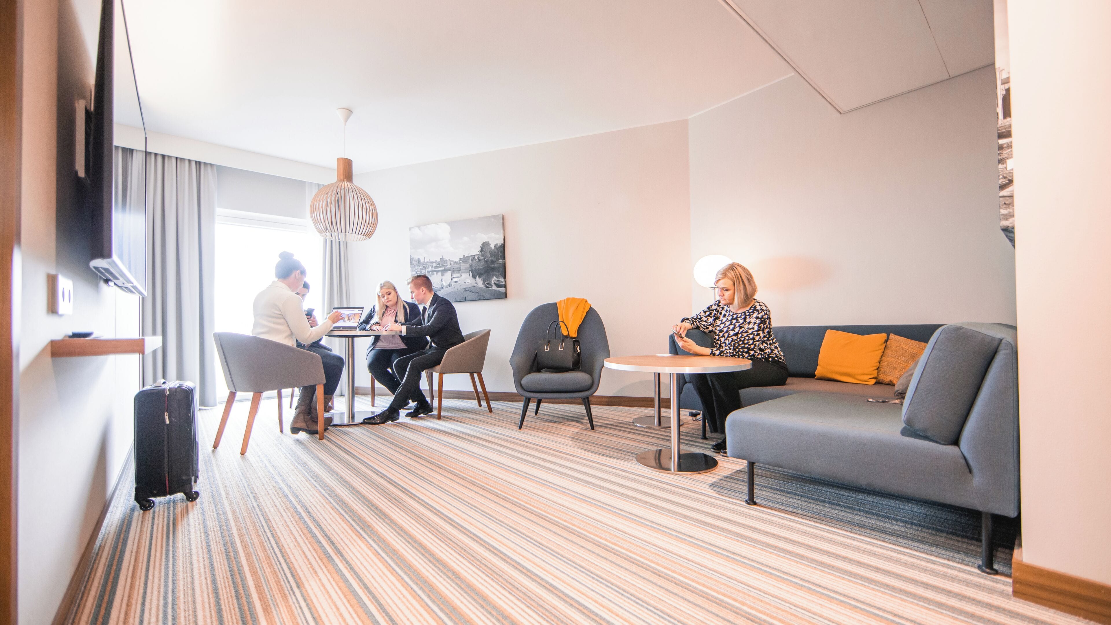 Une chambre d’hôtel avec un tapis à rayures, un canapé gris, une table ronde et une table de réunion autour de laquelle sont assis quatre personnes.