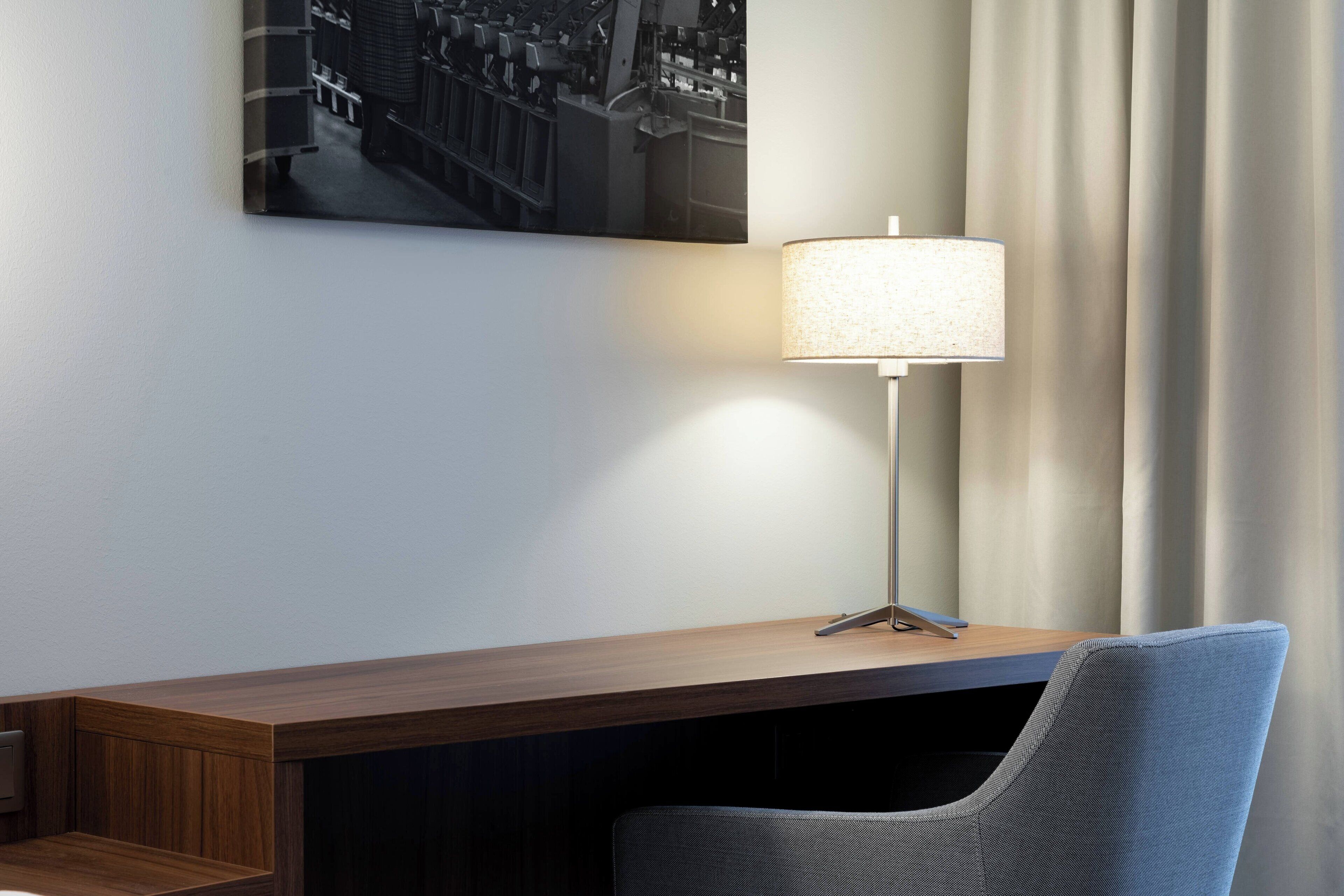 A hotel room with a wooden desk, a modern lamp, a chair, and a cityscape painting on the wall.