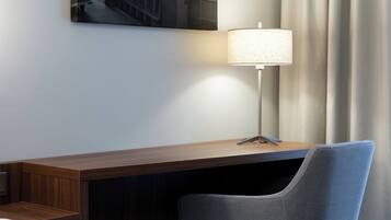 A hotel room with a wooden desk, a modern lamp, a chair, and a cityscape painting on the wall.