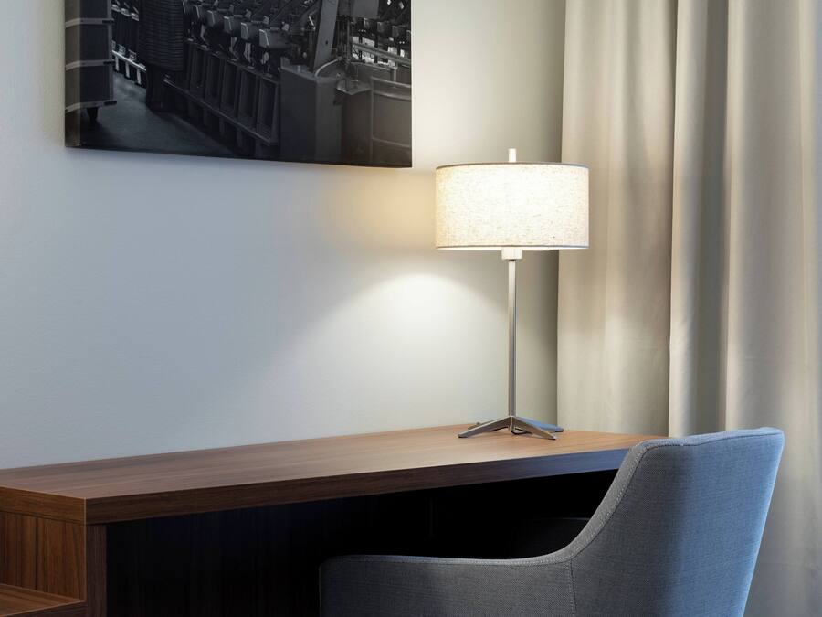 A hotel room with a wooden desk, a modern lamp, a chair, and a cityscape painting on the wall.