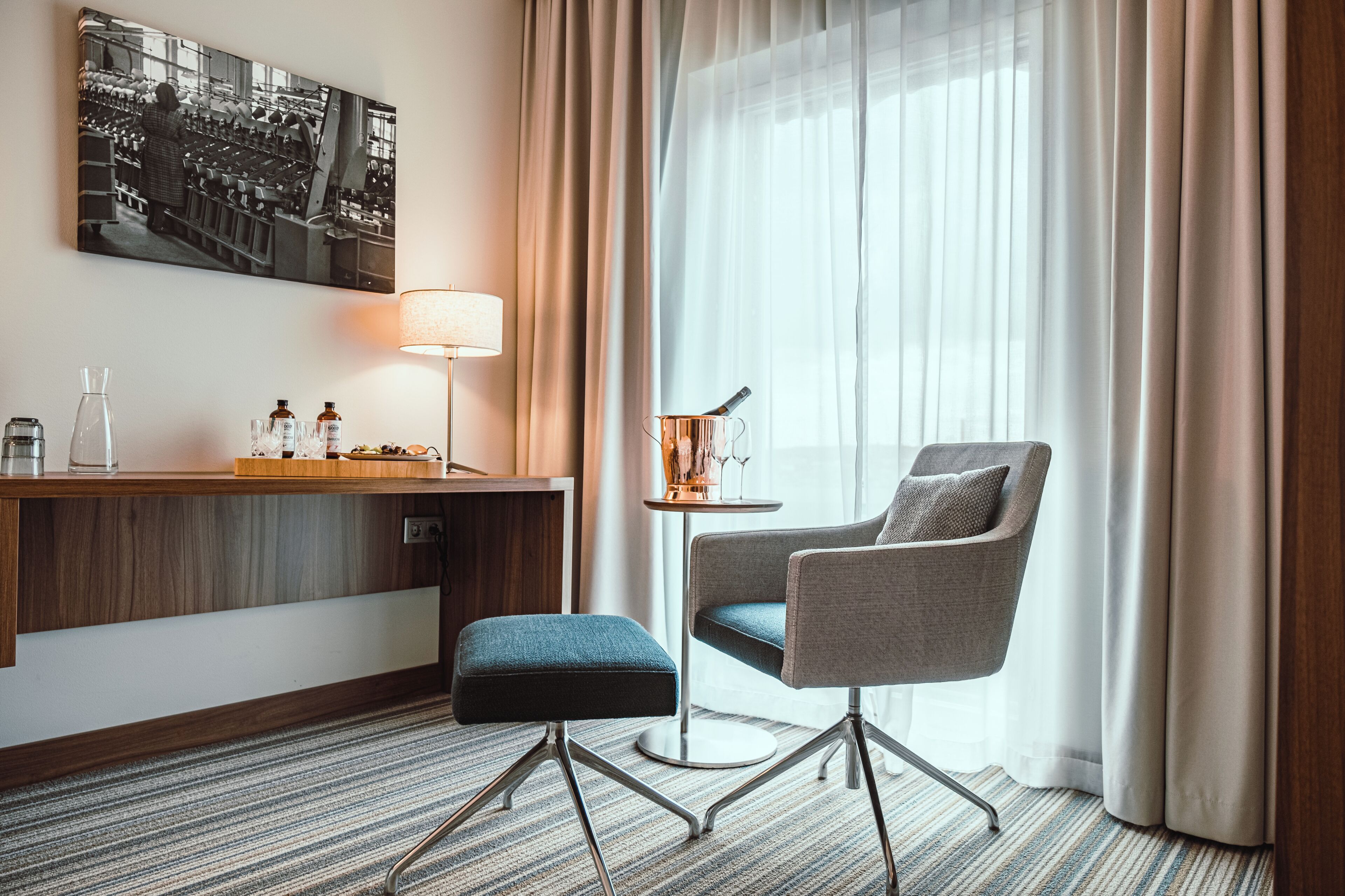 Habitación de hotel con escritorio, silla, lámpara, mesita con una botella y vasos, y una ventana grande con cortinas.