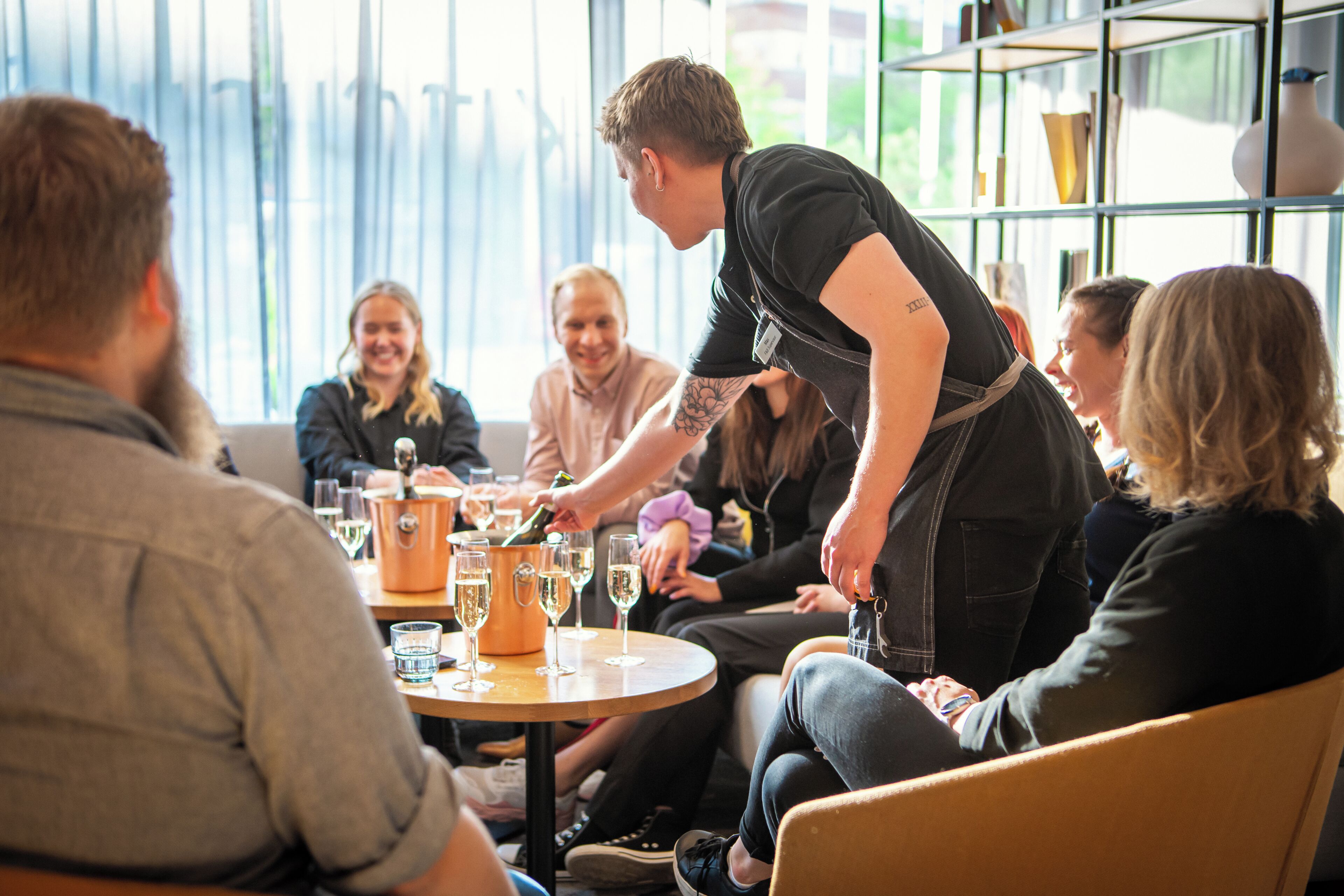 Een groep mensen zit rond een tafel met een fles champagne, glazen en een klein oranje bakje, waarschijnlijk een taartje of dessert.