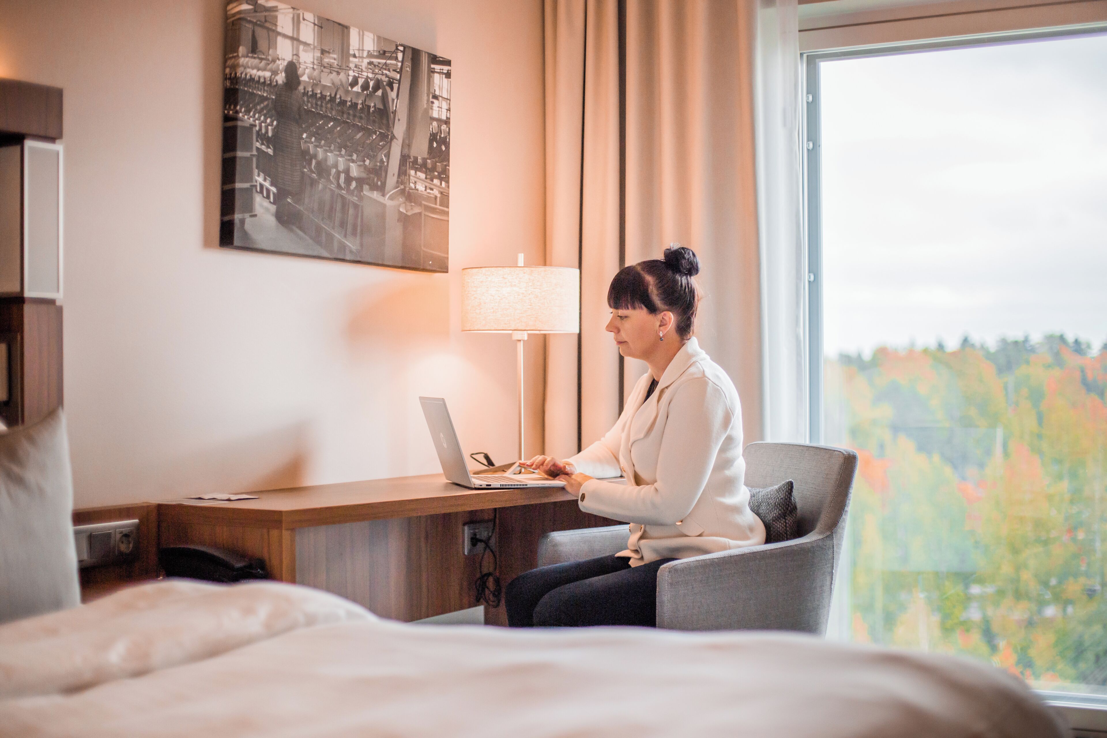 A hotel room with a bed, a desk with a laptop, a lamp, and a window with a view of trees.