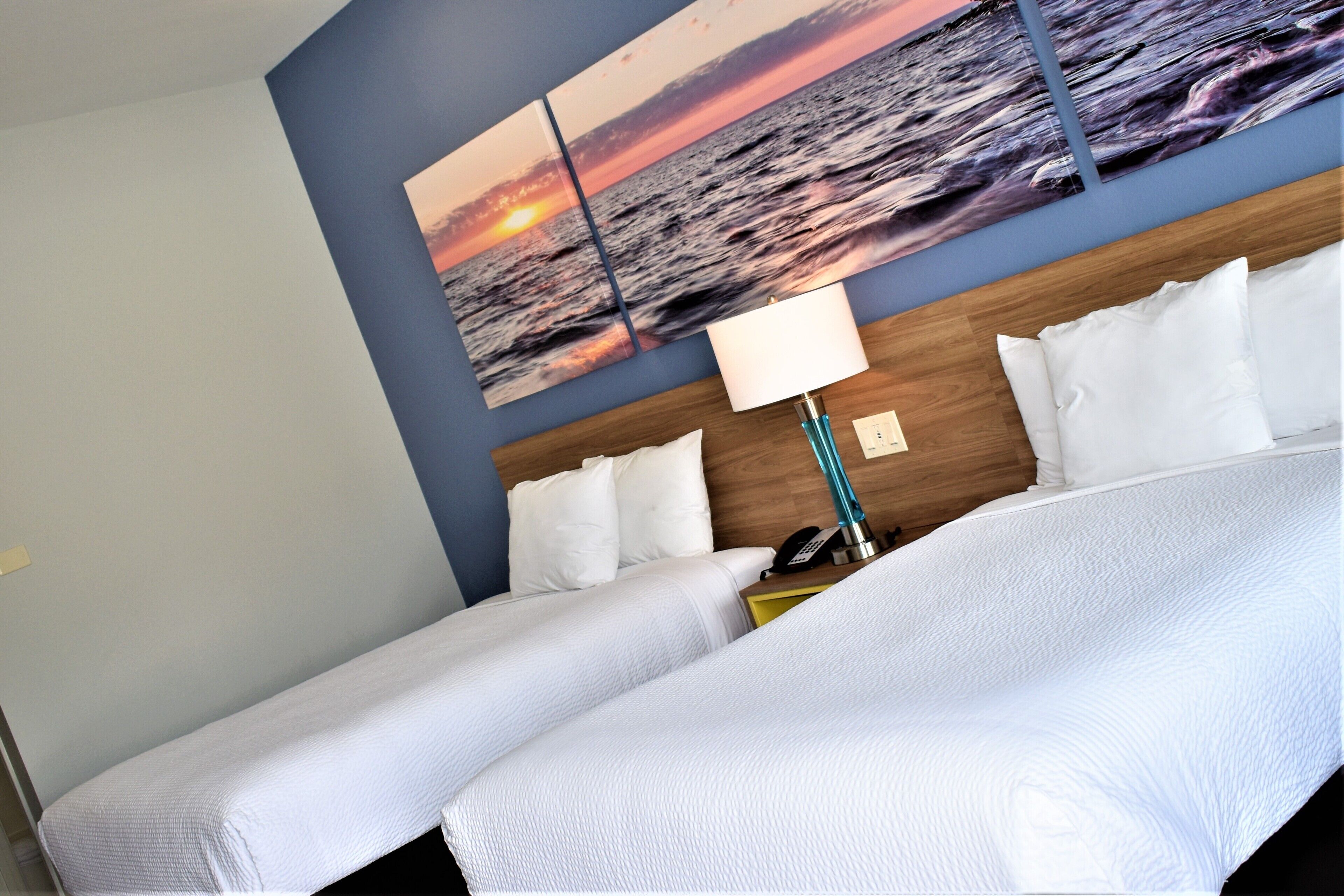 A hotel room with two beds, a nightstand with a lamp, and a wall-mounted painting of a sunset over the ocean.