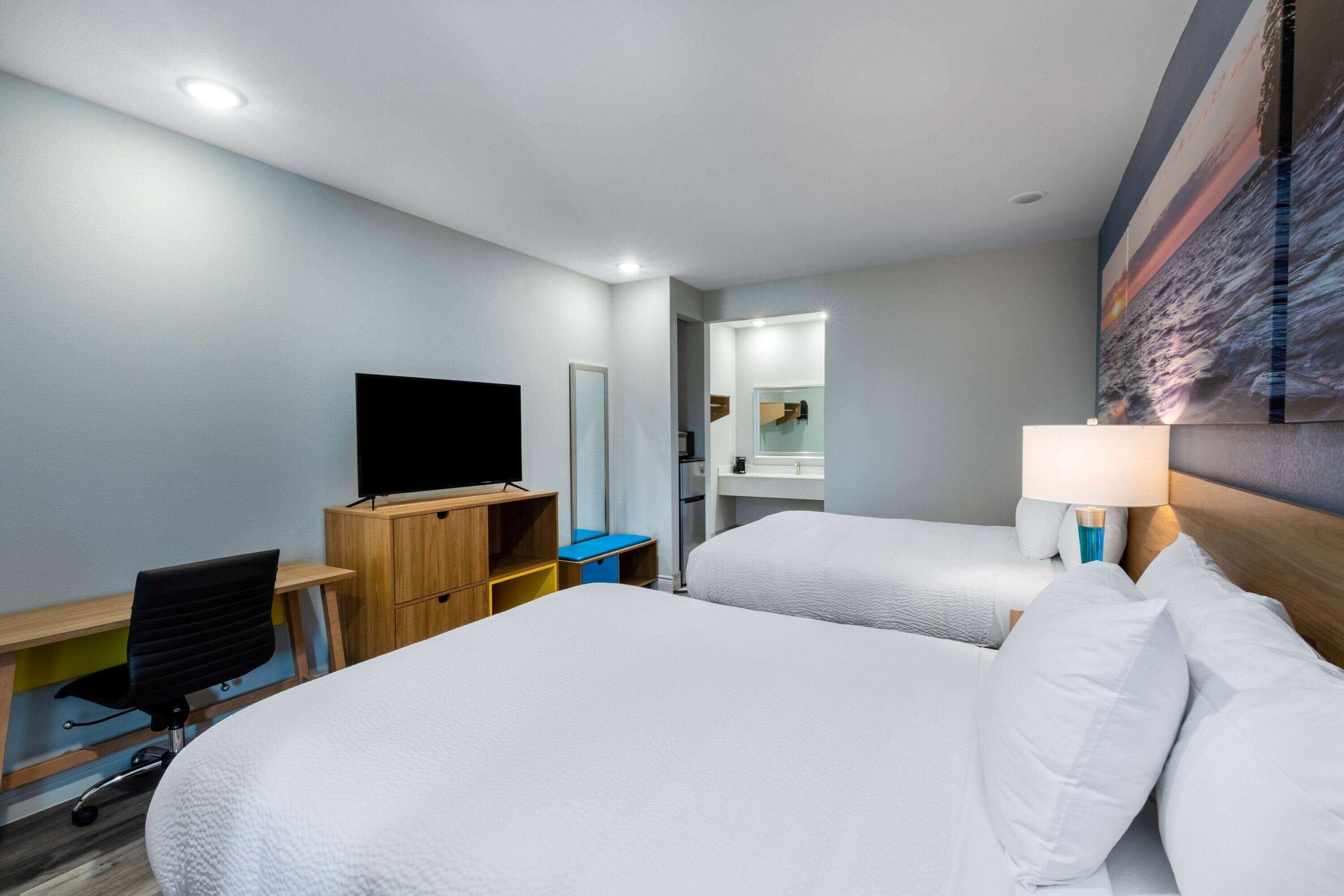 Habitación de hotel con dos camas, un escritorio con silla, televisor, espejo y un cuadro en la pared.