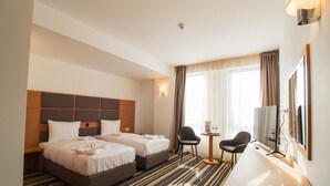 Quarto de hotel com duas camas, uma escrivaninha, cadeiras, televisão e uma janela com cortinas.