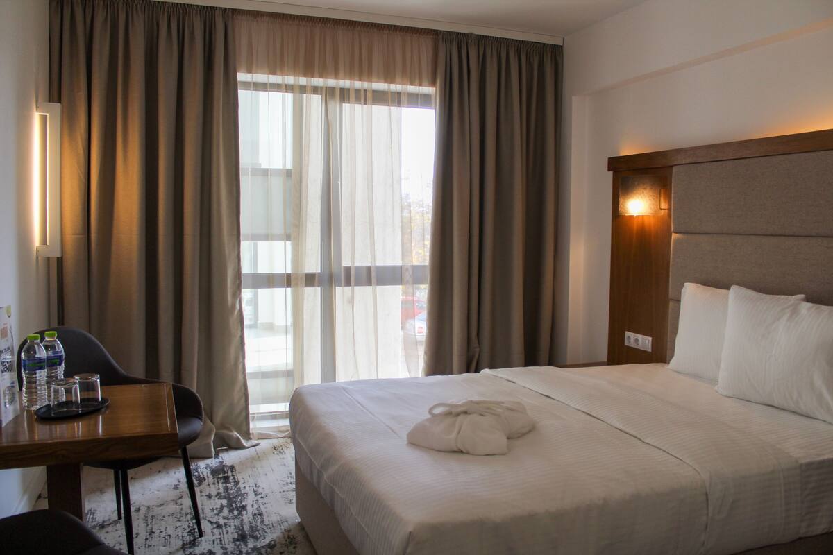 Ein Hotelzimmer mit einem großen Bett, einem Schreibtisch mit Stuhl, einem Tisch mit Wasserflaschen und Blick durch durchsichtige Vorhänge nach draußen.