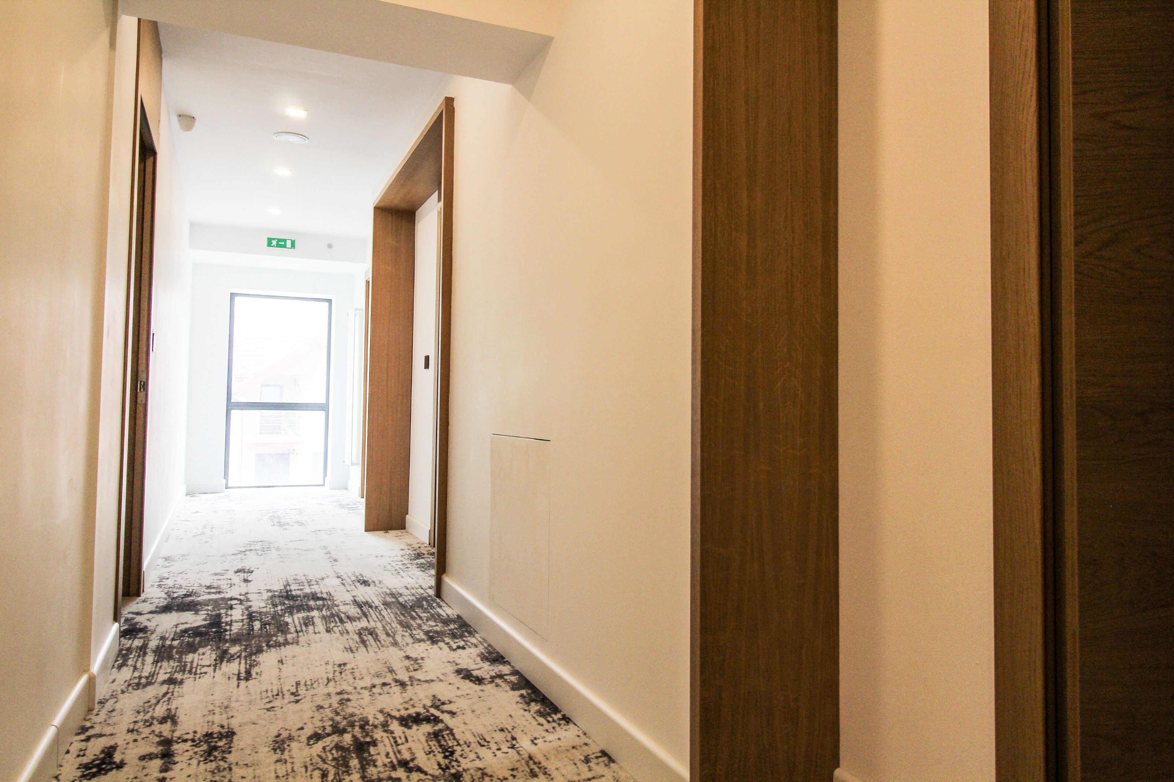 A hotel hallway with a patterned carpet, wooden doors, and a bright exit door at the end.
