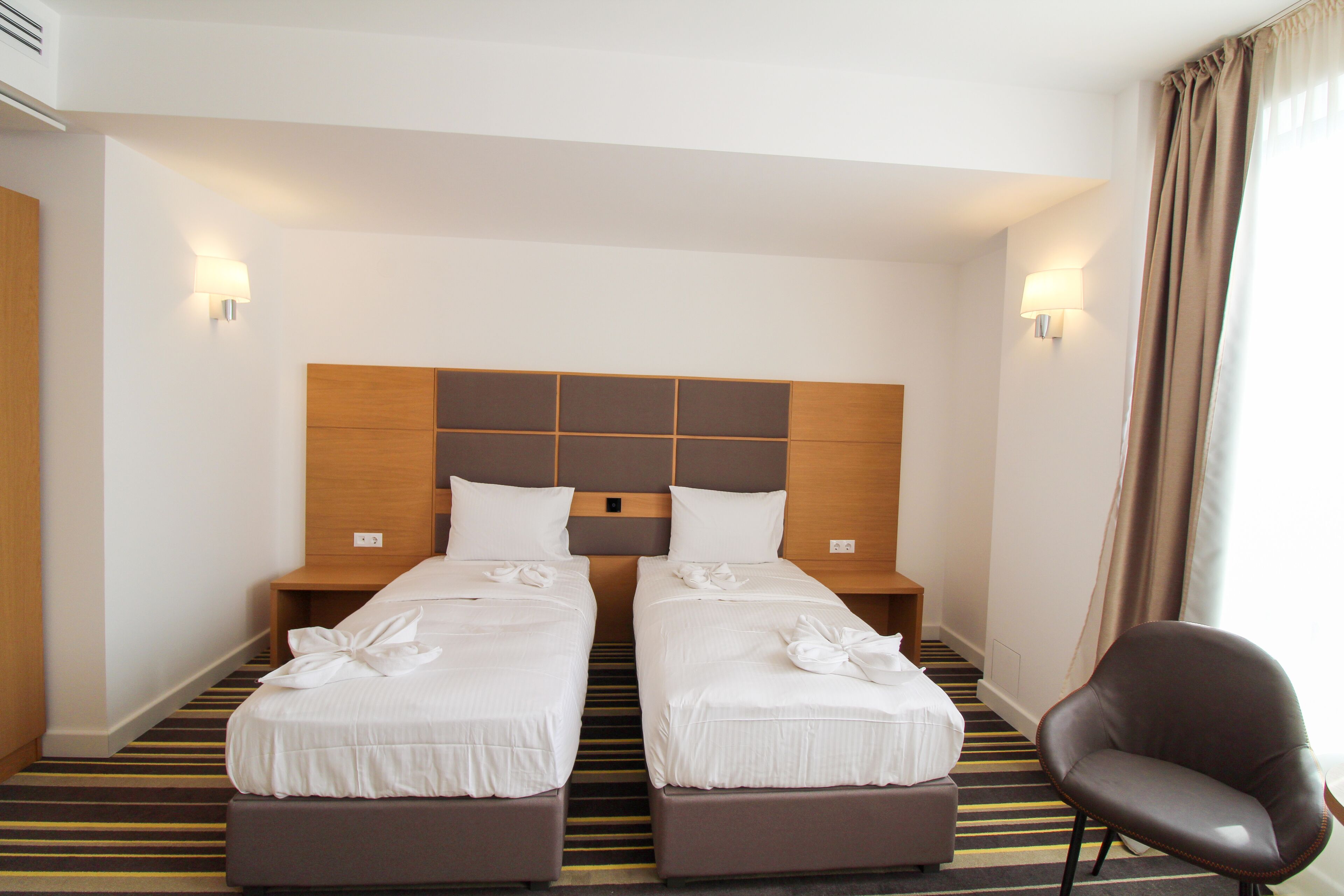 A hotel room with two beds, a wooden headboard, bedside lamps, a striped carpet, and a brown leather chair.
