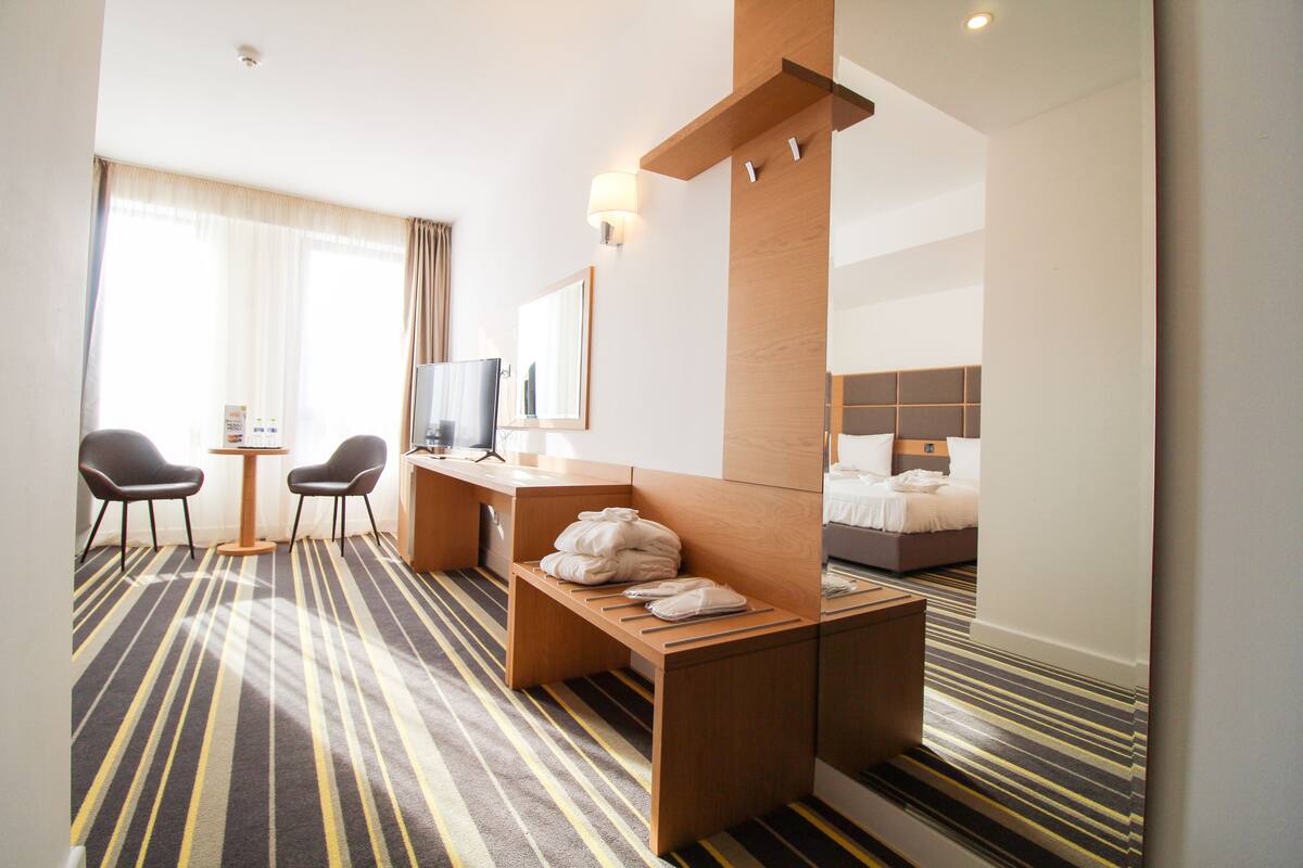 Ein Hotelzimmer mit einem Bett, einem Schreibtisch, zwei Stühlen, einem Fernseher und einem großen Fenster mit Vorhängen.