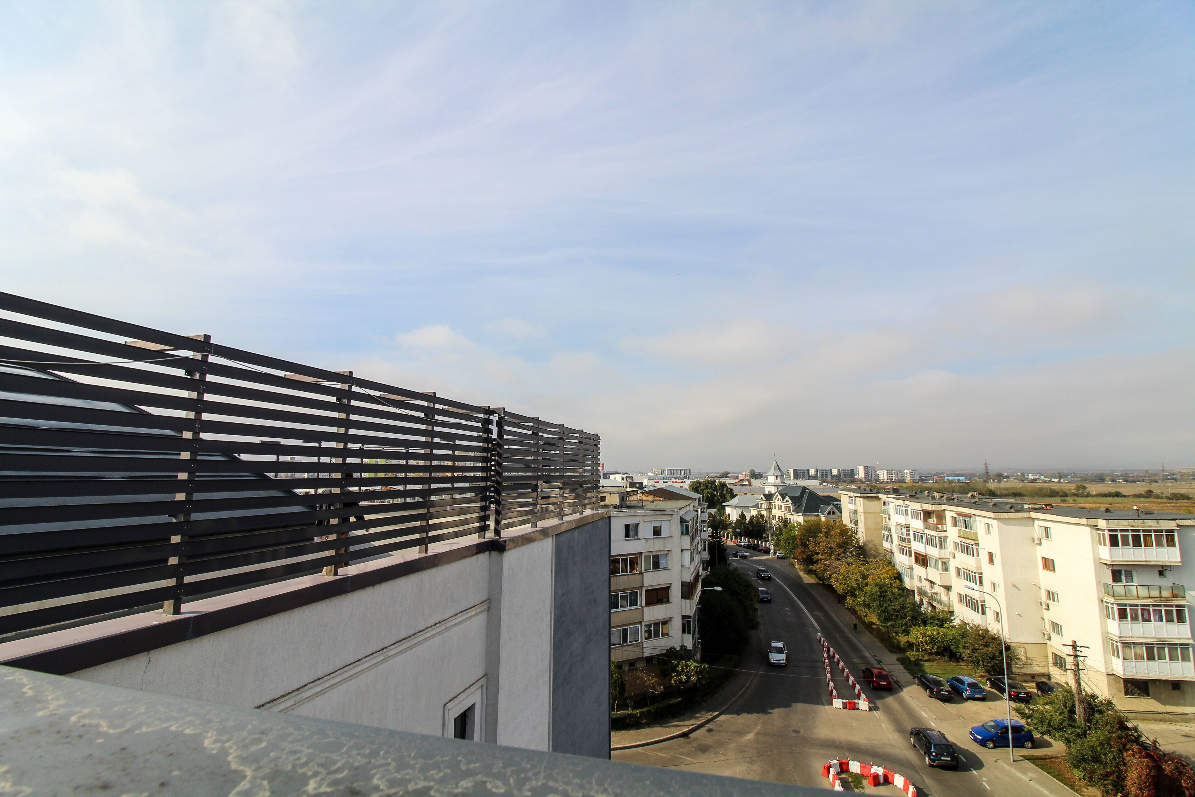 A multi-story building with balconies, a street with parked cars, and a clear sky.