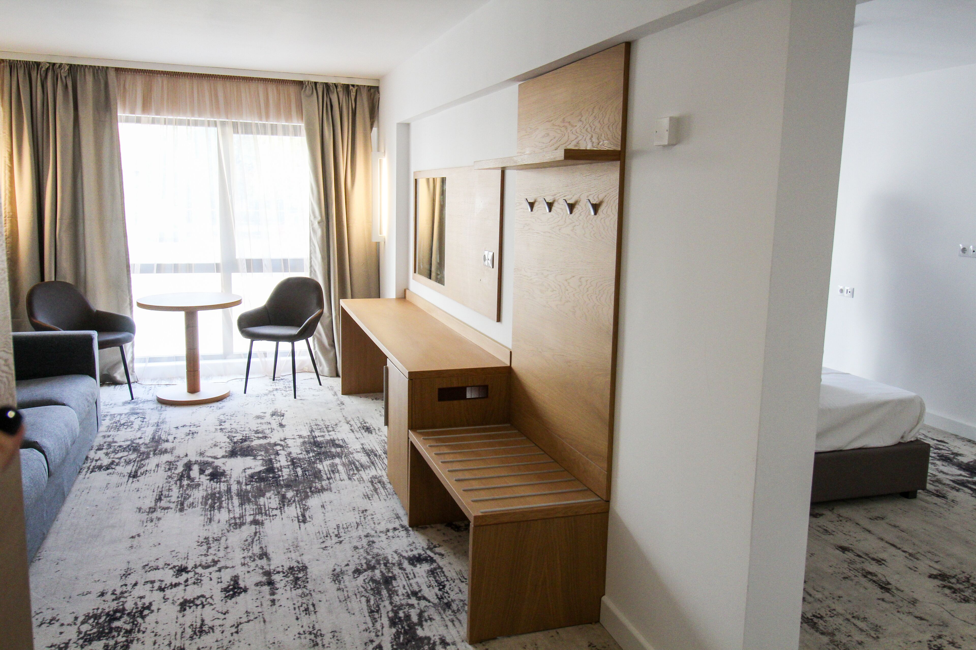 A hotel room with a bed, a desk, two chairs, a small table, and a large window with curtains.