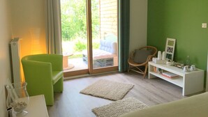 A room with a sliding glass door leading to an outdoor area, a green chair, a white sofa, a white side table with a teapot, and a bookshelf. - Le Jardin des 4M (Saint-Régis-du-Coin)