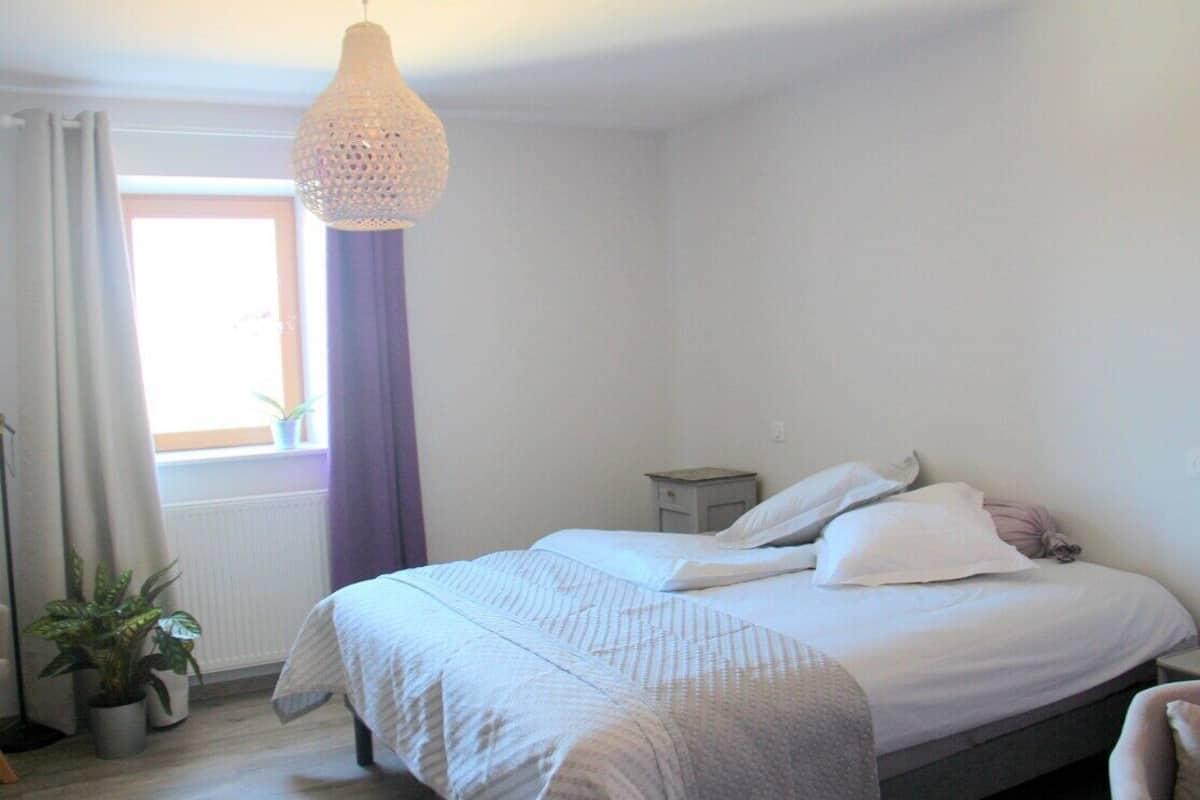 Een slaapkamer met een bed, een nachtkastje, een hanglamp, een raam met gordijnen en een potplant.