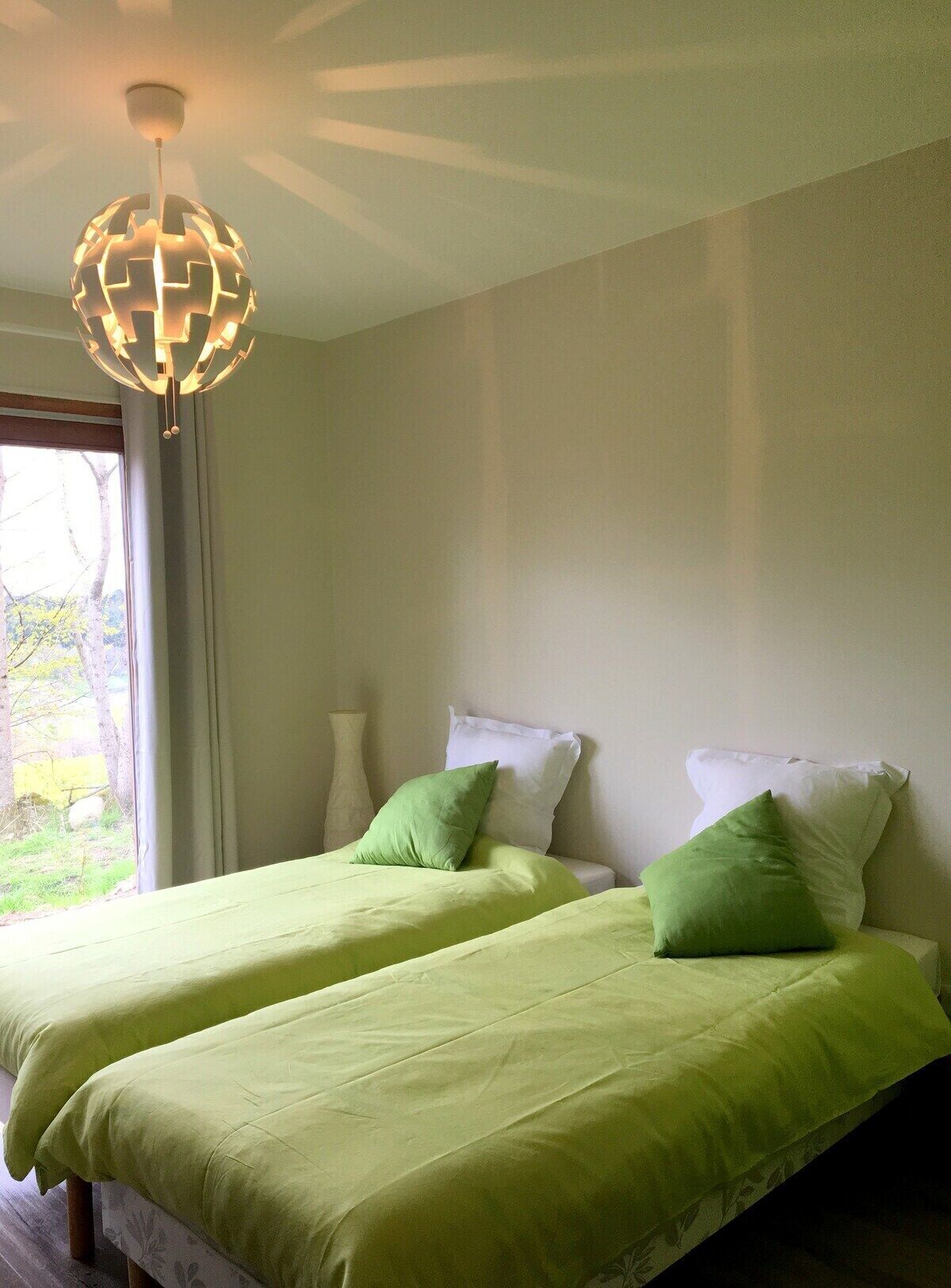 A room with two beds, each with green bedding and white pillows, a modern spherical light fixture, and a window with a view of greenery.