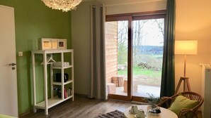A room with a wooden floor, a round table with cups and a lamp, a white shelving unit, and a view through a glass door to an outdoor area with a wooden bench. - Le Jardin des 4M (Saint-Régis-du-Coin)
