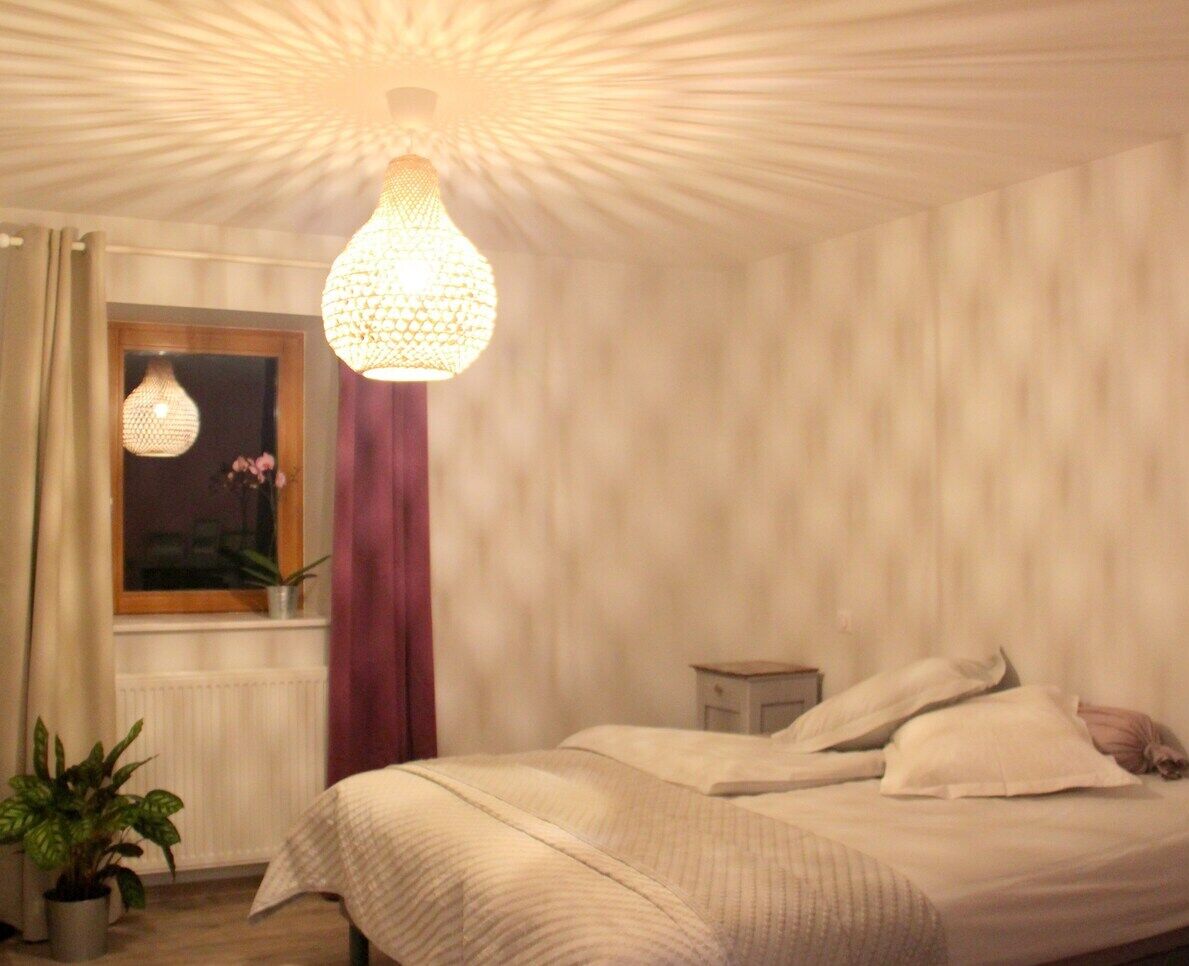 Een slaapkamer met een bed, een raam met gordijnen, een hanglamp en een nachtkastje.