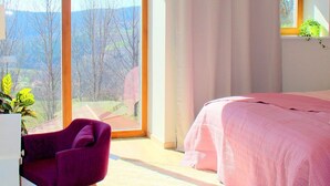 A bedroom with a large window, a bed with pink bedding, a purple chair, and a white desk. - Le Jardin des 4M (Saint-Régis-du-Coin)