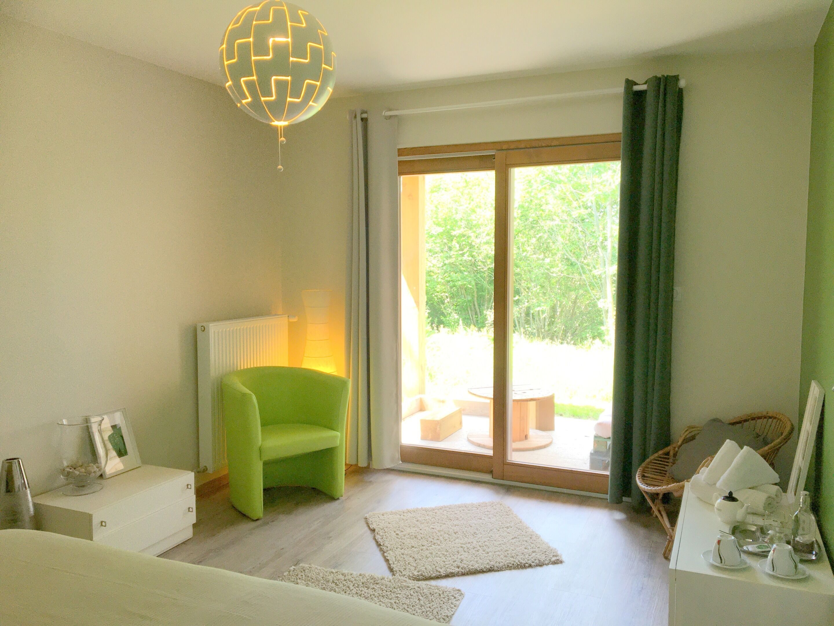 Une pièce avec un fauteuil vert, une commode blanche, un vase en verre et une vue sur la verdure à l’extérieur.
