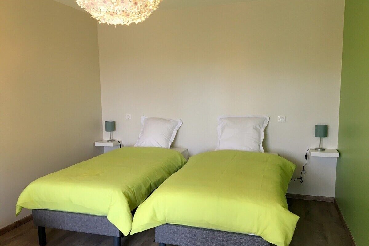 Een kamer met twee eenpersoonsbedden, elk met een groene sprei en witte kussens. Er staan nachtkastjes met lampen bij, en de vloer is van hout.