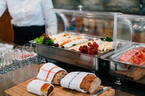 A buffet with various food items, including bread, cheese, grapes, and ham, along with several glasses of water.