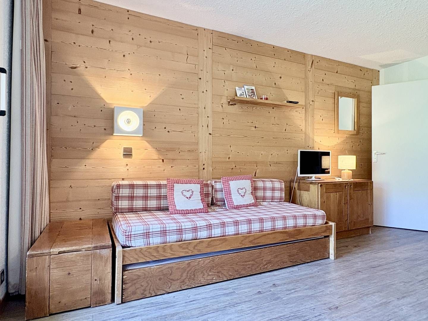 A bedroom with a wooden wall, a bed with plaid bedding, a bedside table with a lamp, and a television.
