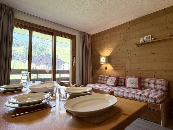 A dining area with a wooden table set for four, a sofa with plaid cushions, and a view of a snowy landscape through large windows.