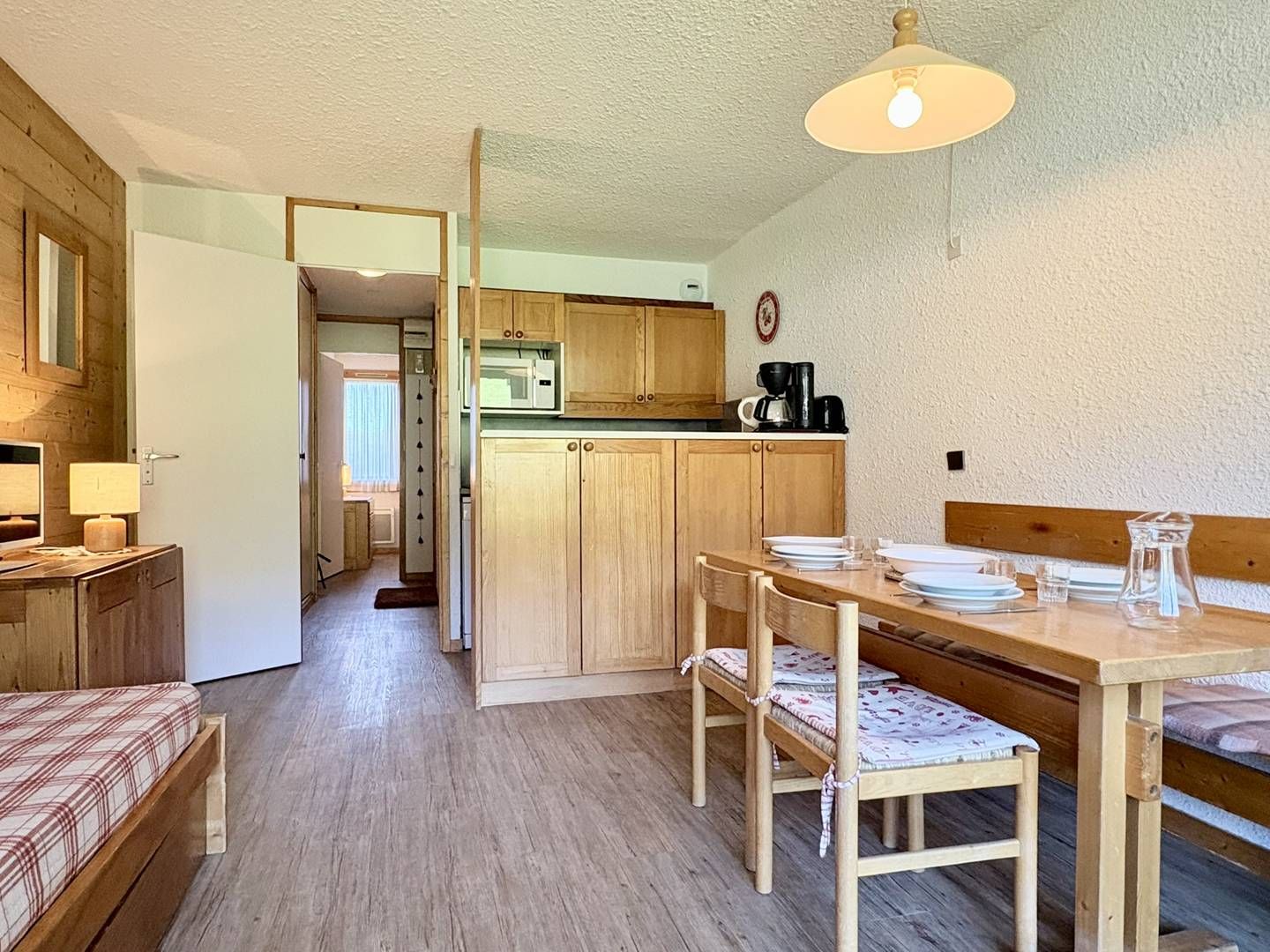 A compact room with a kitchenette, a dining table with chairs, and a bed with a plaid bedspread.