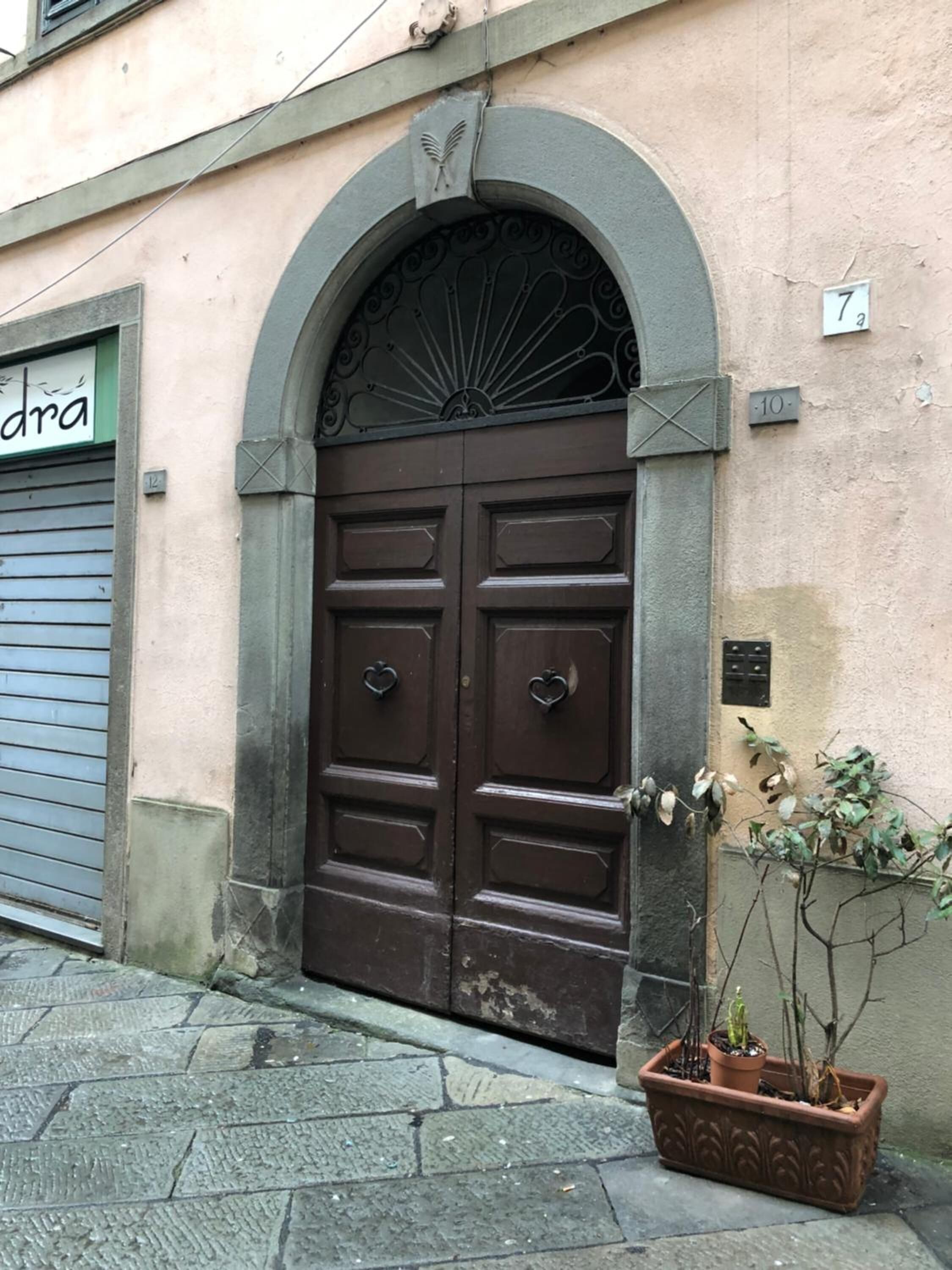 Une porte double marron surmontée d’une arche décorative, un petit pot de fleurs placé à côté de la porte et le numéro de l’immeuble, « 7 », inscrit sur le mur.