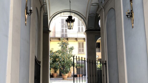 Un pasillo con arcos, un farol colgante y una verja que da a un edificio con balcones.