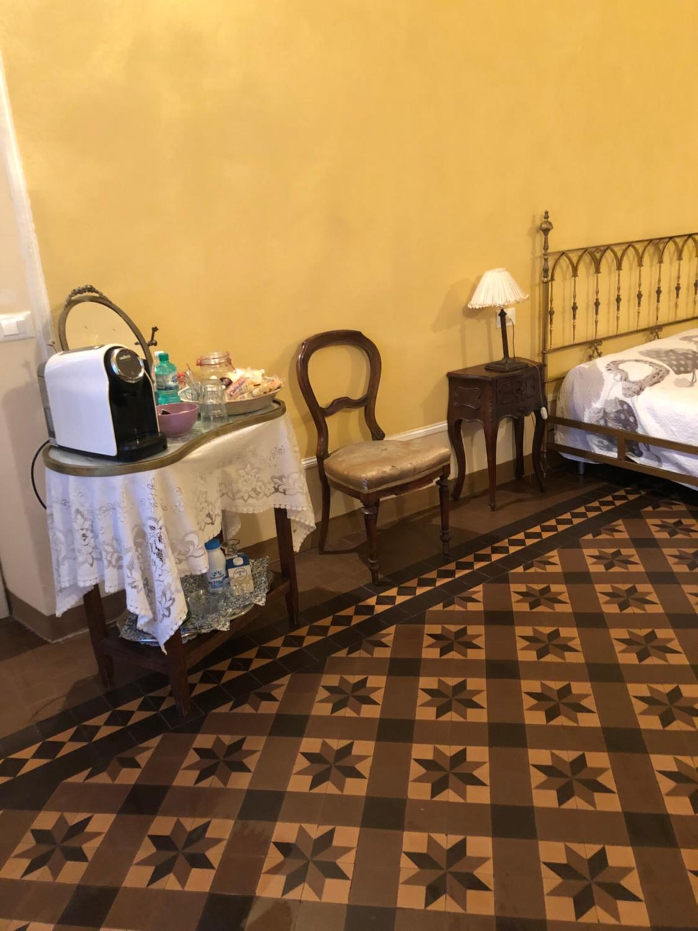 Una stanza con un letto, un tavolo con la macchina del caffè, una sedia e un tavolino con una lampada.